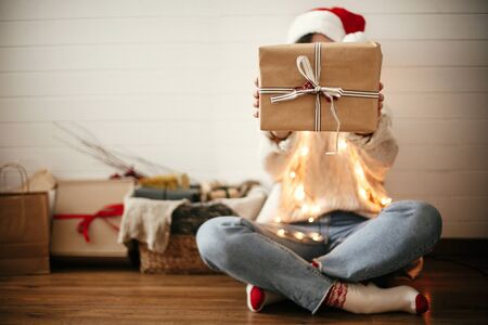 Simple christmas gift box with ribbon in hands of stylish happy girl in santa hat on background of modern christmas tree, lights and stockings. Merry Christmas and Happy holidays. Copy Spaceの写真素材