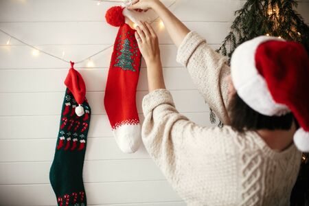 Stylish girl in cozy sweater and santa hat decorating room for christmas holidays with stockings, garland light and christmas tree on white wall . Merry Christmas. Happy Holidaysの写真素材