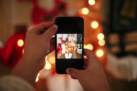 Hands holding phone and taking photo of christmas gift boxes, santa hat, lights on wooden background...