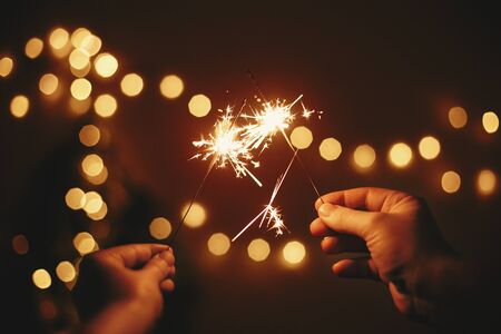 Glowing sparklers in hands on background of golden christmas tree lights, couple celebrating in dark festive room. Happy New Year. Space for text. Fireworks burning in hands. Happy Holidaysの写真素材