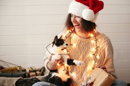 Stylish happy girl in santa hat playing with cute cat in festive christmas lights on background of modern room with presents. Young hipster woman in cozy sweater hugging kitty and smilingの写真素材
