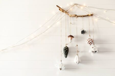 Christmas vintage toys hanging on wooden branch on white wall with festive lights in modern room, scandinavian minimal style. Stylish glass ornaments, holiday decorations. Space for textの写真素材