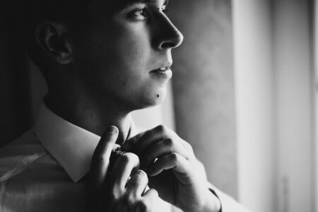 Stylish groom in white shirt putting on bow tie near window in hotel room. Morning preparation before wedding ceremony. Man getting ready before luxury eventの写真素材