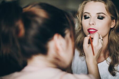 Beautiful happy bride getting her makeup done by professional make up artist in hotel room. Morning preparation before wedding ceremony. Stylist applying red lipstickの写真素材