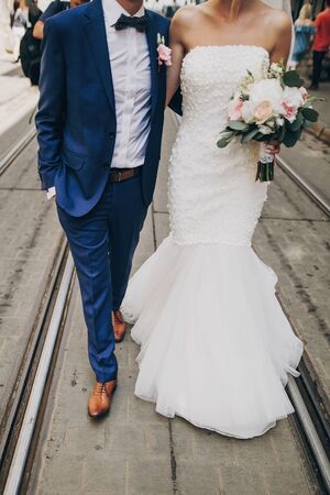 Stylish bride with bouquet and groom walking in sunny european city street, cropped view. Gorgeous wedding couple of newlyweds embracing outdoors. Romantic moment