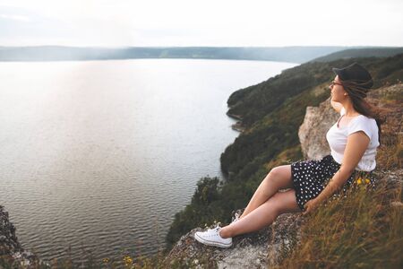 Traveler hipster girl in hat and windy hair sitting on top of rock mountain with wildflowers, enjoying beautiful sunset on river. Calm tranquil moment. Copy spaceの写真素材