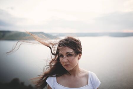 Portrait of hipster girl with windy hair standing on top of rock mountain with beautiful sunset view on river. Atmospheric tranquil and calm moment. Copy space.の写真素材