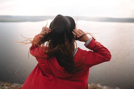 Traveler girl in hat and with windy hair standing in evening sunlight on top of rock mountain, enjoying beautiful sunset view on river. Back of woman in red jacket relaxing on cliff. Copy spaceの写真素材