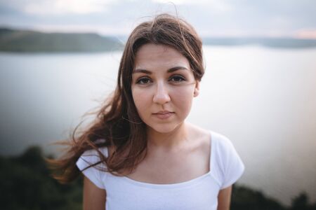 Traveler girl with windy hair relaxing on top of rock mountain, enjoying beautiful sunset view on river. Atmospheric moment. Copy space. Calm portrait of brunette woman in white shirtの写真素材