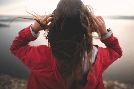 Traveler girl with windy hair holding hat and looking at evening sky on top of rock mountain, enjoying beautiful sunset view on river. Copy space. Atmospheric momentの写真素材