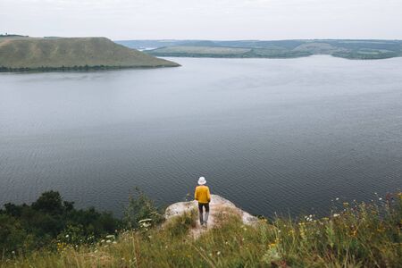 Hipster teenager standing on top of rock mountain and looking on river, view from back. Young stylish guy exploring and traveling. Atmospheric tranquil moment. Copy spaceの写真素材