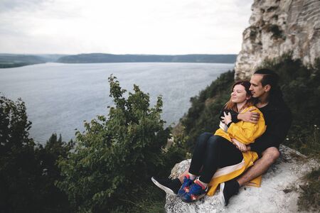 Happy hipster couple in raincoats sitting on top of rock mountain with beautiful view on river. Tourist couple hugging and sitting on windy cliff. Travel and wanderlust. Copy spaceの写真素材