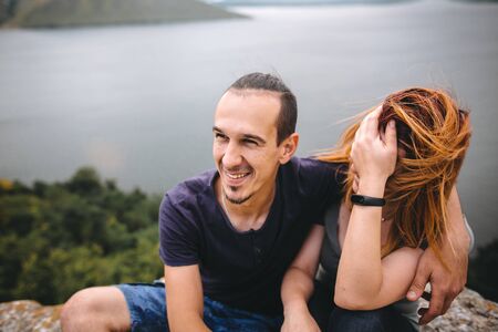 Happy hipster couple sitting and laughing on top of rock mountain with beautiful view on river. Tourist couple hugging and smiling on windy cliff. Traveling together. Copy spaceの写真素材
