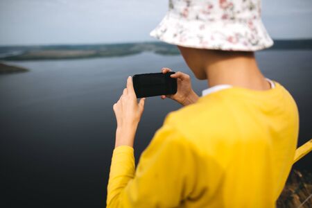Hipster teenager taking photo on phone of amazing view on river while standing on top of rock mountain. Young stylish guy exploring and traveling. Atmospheric moment. Copy spaceの写真素材