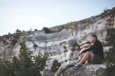 Hipster traveler sitting on top of rock mountain and enjoying amazing view on river. Stylish guy in windbreaker exploring and traveling. Atmospheric tranquil moment. Copy spaceの写真素材
