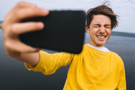 Stylish hipster teenager taking selfie on phone and smiling on top of rock mountain with amazing view on river. Young guy in yellow pullover exploring and traveling. Copy spaceの写真素材