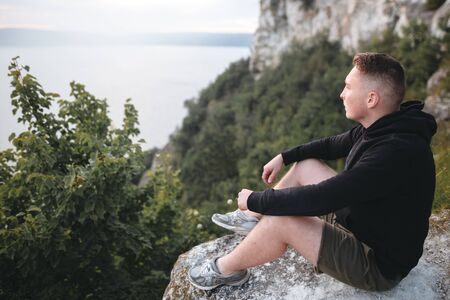 Hipster traveler sitting on top of rock mountain and enjoying amazing view on river. Stylish guy in windbreaker exploring and traveling. Atmospheric tranquil moment. Copy spaceの写真素材