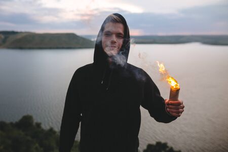 Ultras hooligan holding sparkler with burning fire in hand while standing on top of rock mountain with amazing sunset view on river . Atmospheric moment. Copy spaceの写真素材