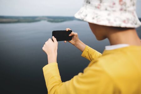 Hipster teenager taking photo on phone of amazing view on river while standing on top of rock mountain. Young stylish guy exploring and traveling. Atmospheric moment. Copy spaceの写真素材