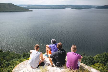 Group of hipster travelers sitting on top of rock mountain and enjoying  amazing view on river. Young guys explorers relaxing and traveling. Atmospheric tranquil moment. Copy spaceの写真素材