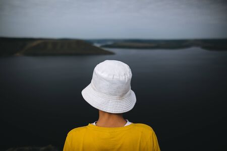 Hipster teenager in yellow pullover standing on top of rock mountain and looking on river, view from back. Young stylish guy exploring and traveling. Atmospheric tranquil momentの写真素材