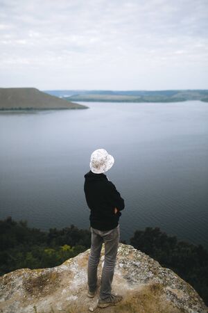 Hipster teenager standing on top of rock mountain and looking on river, view from back. Young stylish guy exploring and traveling. Atmospheric tranquil moment. Copy spaceの写真素材