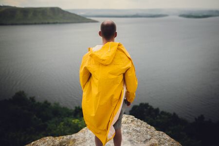 Hipster traveler in yellow raincoat standing on cliff and looking at lake in windy moody day. Wanderlust and travel concept. Man hiking in Norway on foggy day. Atmospheric momentの写真素材