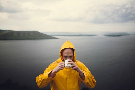 Hipster traveler in yellow raincoat holding metal mug and standing on cliff in rainy windy day with view on lake. Wanderlust and travel. Brutal bearded man hiking. Atmospheric momentの写真素材