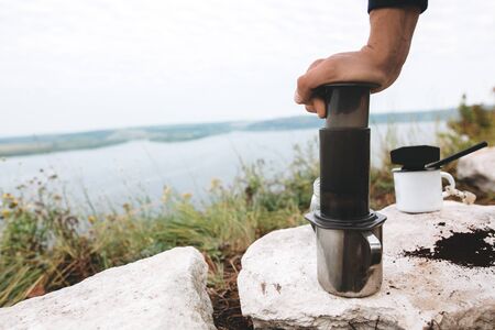 Traveler pressing aeropress on metal mug on cliff at lake, brewing alternative coffee at camping. Making hot drink at picnic outdoors. Trekking and hiking in mountainsの写真素材