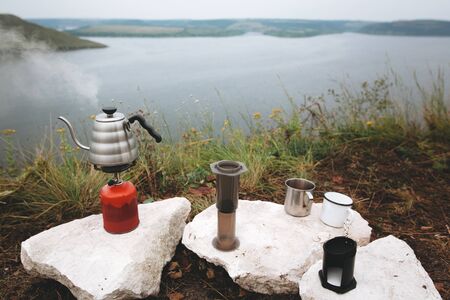 Steel kettle boiling on gas primus, metal mugs and aeropress on cliff on background of lake, brewing coffee at camping. Making hot drink at picnic outdoors. Trekking and hiking in mountainsの写真素材