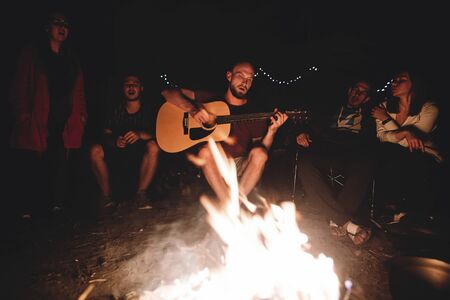 Hipster man playing on acoustic guitar and singing song with friends travelers at big bonfire at...