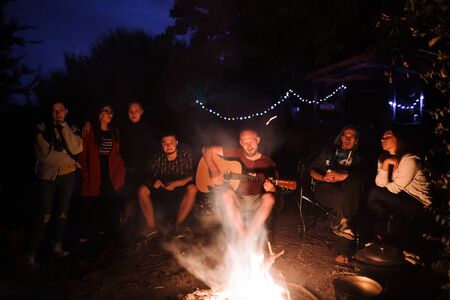 Friends travelers chilling at big bonfire, listening to acoustic music, relaxing at camp in the night forest. Group of people resting at fire in the evening, camping near lakeの写真素材