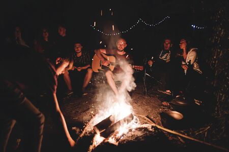 Hipster man playing on acoustic guitar and singing song with friends travelers at big bonfire at night camp in the forest. Group of people chilling at fire in the evening, camping near lakeの写真素材