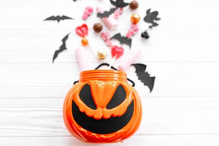 Halloween candy with skulls, black bats, ghost, spider decorations spilled from jack o lantern bucket on white woodenの写真素材