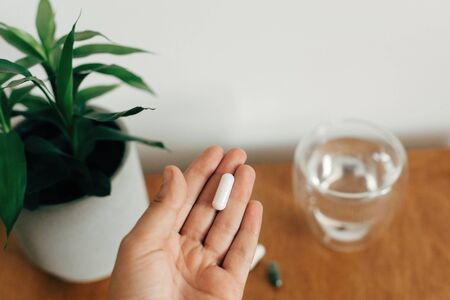 Hand holding magnesium capsule above glass of water on wooden table. White pill. Dietary supplements. Health support and treatment. Biologically active additivesの写真素材