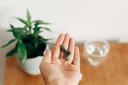 Hand holding spirulina capsule above glass of water on wooden table. Morning vitamin nutrient pill. Dietary supplements. Health support and treatment. Biologically active additivesの写真素材