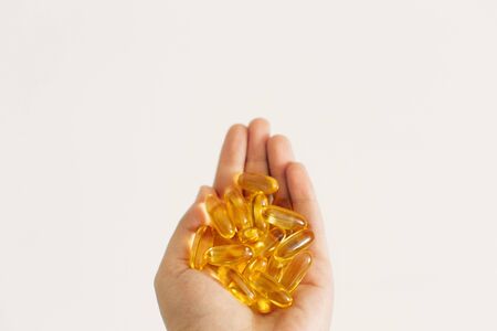 Hand holding omega 3 capsules on white wall background. Morning dose of vitamin. Fish oil in pills. Health support and treatment. Dietary supplements. Biologically active additivesの写真素材