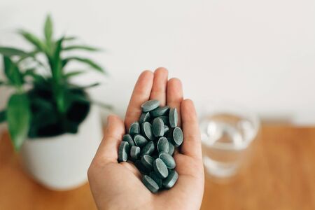 Hand holding chlorophyll tablets above glass of water on wooden table. Morning vitamin nutrient pill. Dietary supplements. Health support and treatment. Biologically active additivesの写真素材
