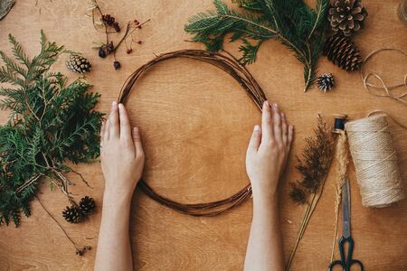 Making christmas wreath. Hands holding wooden circle and fir branches, pine cones, thread, scissors, herbs on wooden table. Details for workshop of making christmas wreathの写真素材