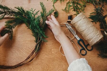 Hands holding fir branches and pine cones, thread, scissors on wooden table. Making rustic christmas...