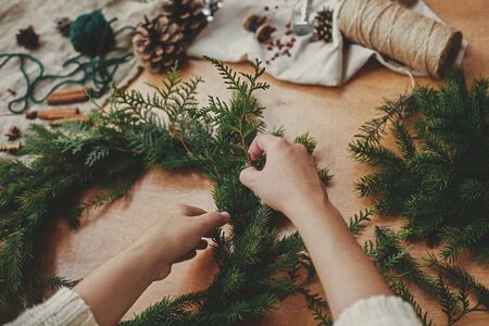 Hands holding fir branches, and pine cones, thread, berries, scissors on wooden table. Christmas wreath workshop. Authentic stylish still life. Making rustic Christmas wreath.の写真素材