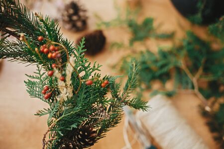 Rustic Christmas wreath. Close Up of berries and fir branches on christmas wreath with pine cones and herbs, scissors and thread on rural wooden table. Happy holidays. Workshopの写真素材