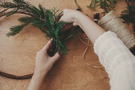 Making rustic christmas wreath flat lay. Hands holding fir branches and pine cones, thread, scissors on wooden table. Details for workshop of making christmas wreathの写真素材