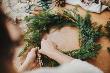 Making rustic Christmas wreath. Hands holding herbs and fir branches, pine cones, thread, berries, cinnamon on wooden table. Christmas wreath workshop. Authentic stylish still life.の写真素材