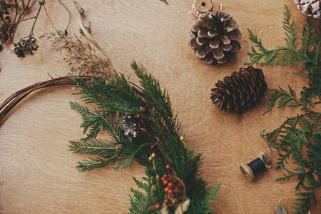 Rustic Christmas wreath, flat lay. Creative christmas wreath with fir branches, berries, pine cones and herbs with thread on rural wooden table. Happy holidays. Workshopの写真素材