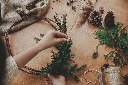 Christmas wreath workshop. Hands holding herbs, fir branches, pine cones, berries, thread, scissors on wooden table. Making rustic christmas wreath. Authentic rural wreathの写真素材