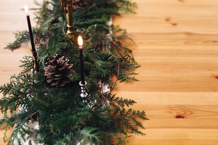 Stylish rustic christmas arrangement for festive dinner. Fir branches with pine cones and vintage candlestick with burning black candles on wooden table. Christmas rural table decor for feastの写真素材