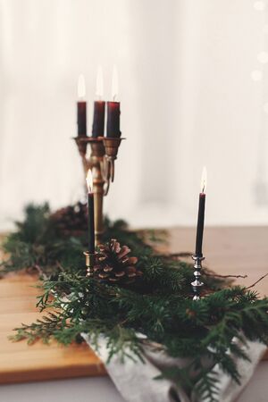 Stylish rustic christmas arrangement for festive dinner. Fir branches with pine cones and vintage candlestick with burning black candles on wooden table. Christmas rural table decor for feastの写真素材