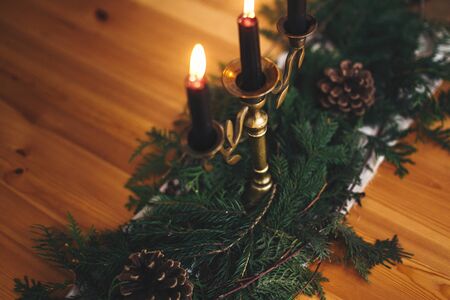 Fir branches with pine cones and vintage candlestick with burning black candles on wooden table. Stylish rustic christmas arrangement for festive dinner. Christmas rural table decor for feastの写真素材