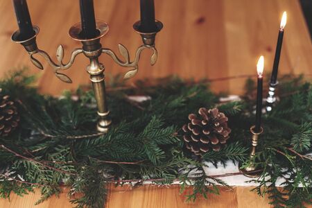 Fir branches with pine cones and vintage candlestick with burning black candles on wooden table. Stylish rustic christmas arrangement for festive dinner. Christmas rural table decor for feastの写真素材